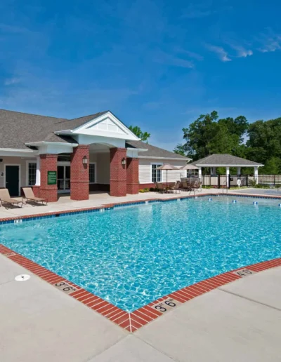 A large outdoor swimming pool with brick edging and a sundeck located next to a traditional clubhouse under a clear blue sky