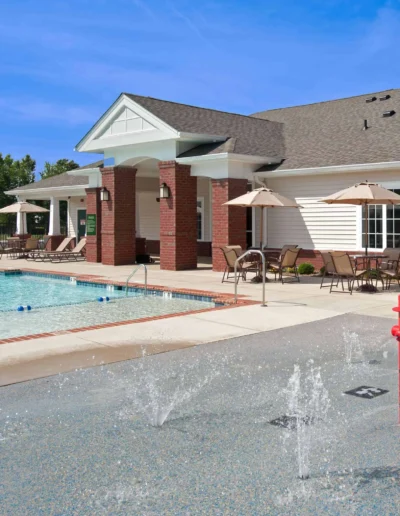 An outdoor children's splash pad area featuring water jets and a red fire hydrant sprayer next to the community swimming pool and clubhouse